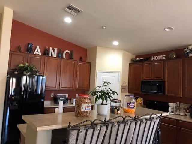 Open kitchen with a central island and bar stools, dark wood cabinets, black refrigerator and microwave, countertop jars and a potted plant.