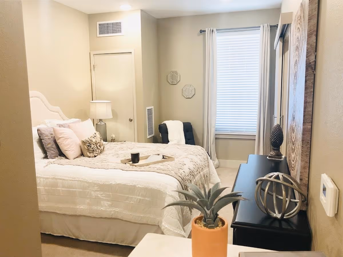 A cozy bedroom with a neatly made bed featuring white and beige bedding and multiple pillows. A bedside table with a lamp is next to the bed. There is a window with white blinds and curtains, a black chair with a white throw blanket, and a black dresser with decorative items including a spherical sculpture and a pinecone-shaped ornament. A small potted plant is visible in the foreground.