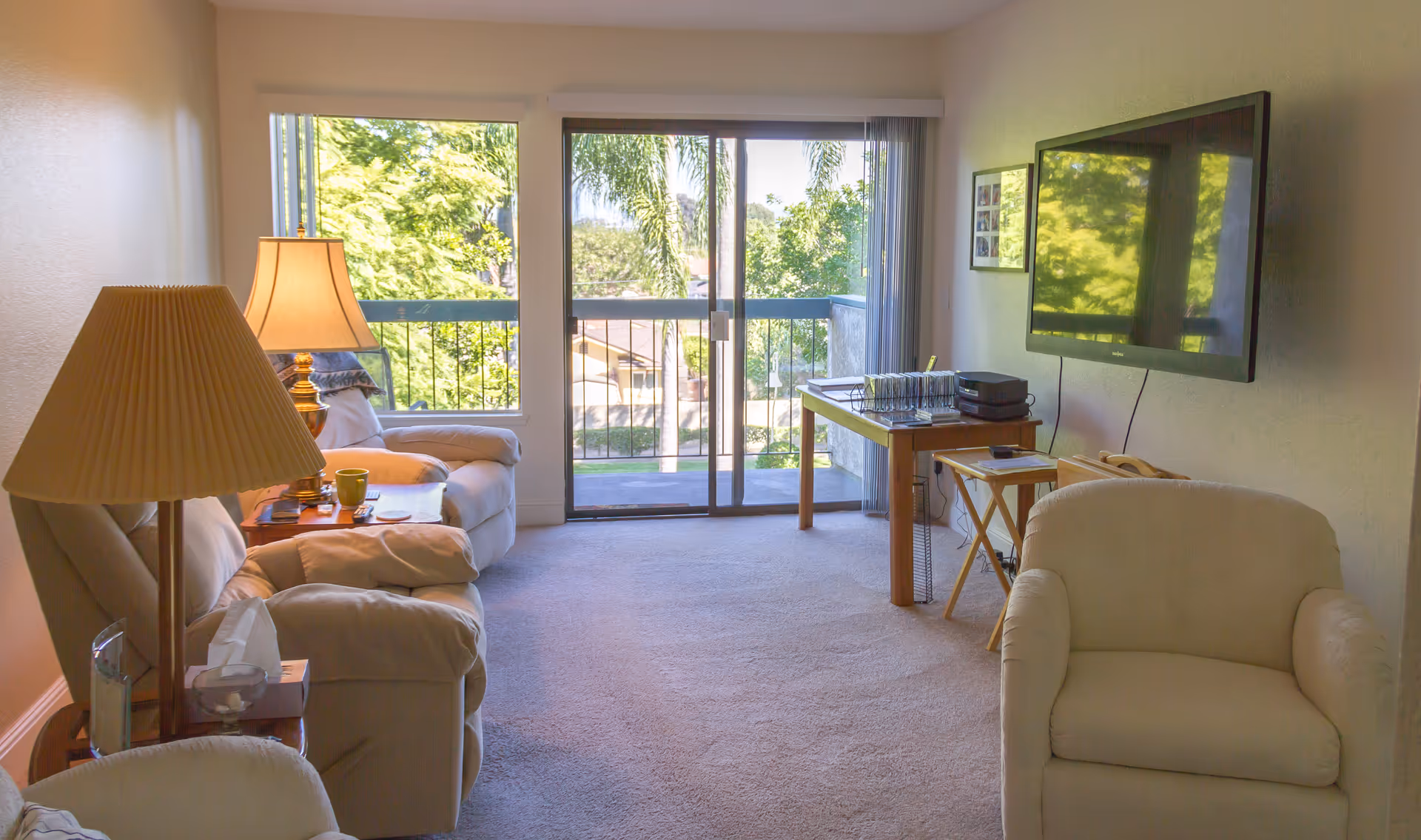 A cozy living room with beige armchairs and a sofa, a wooden side table with a lamp and a cup, a wall-mounted flat-screen TV, and a wooden table with office supplies near a sliding glass door that opens to a balcony with a view of trees and greenery outside.