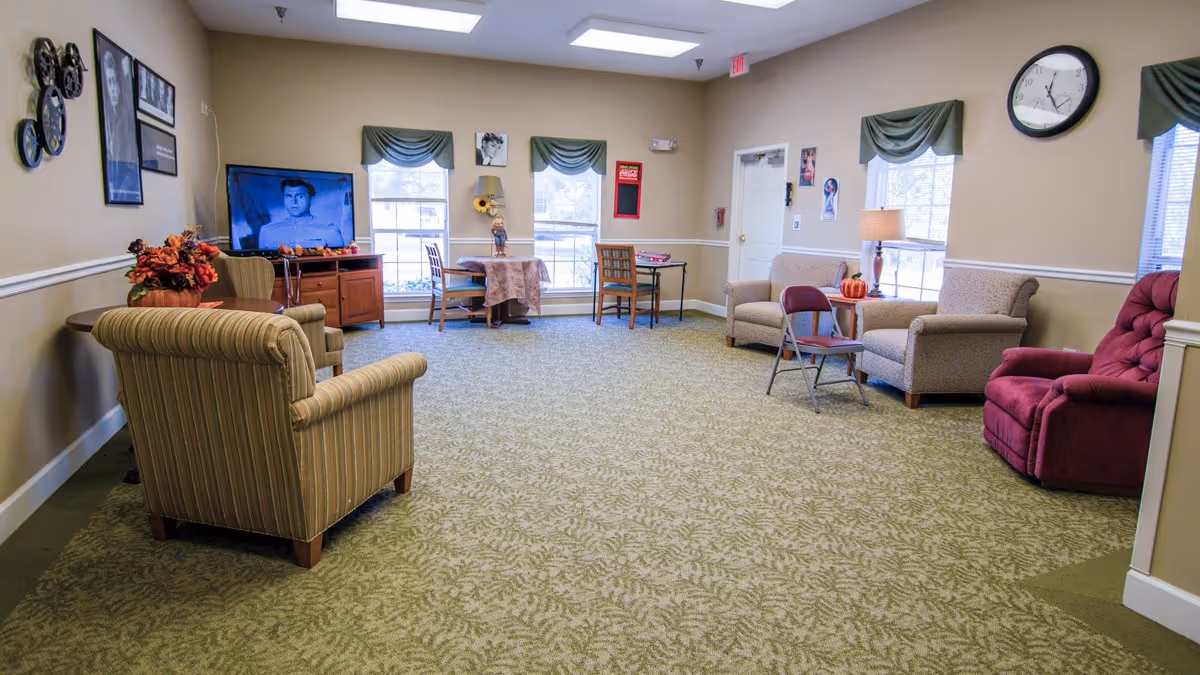 A cozy living room area in Brookdale Hampton Cove featuring various seating options including armchairs, a recliner, and a folding chair. The room has beige walls with white trim, green patterned carpet, and three windows with green valances. A TV is on a wooden stand showing a black and white image. There are small tables with decorative items, a wall clock, and framed pictures on the walls.
