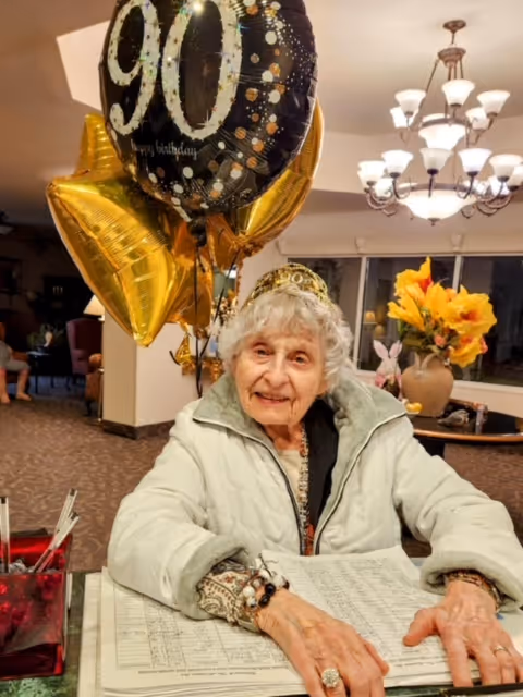 An elderly woman wearing a light-colored jacket and a tiara is sitting at a table with a large open book in front of her. Behind her are gold star-shaped balloons and a large black balloon with '90' and 'happy birthday' written on it. The background shows a chandelier, a vase with yellow flowers, and a cozy indoor setting.