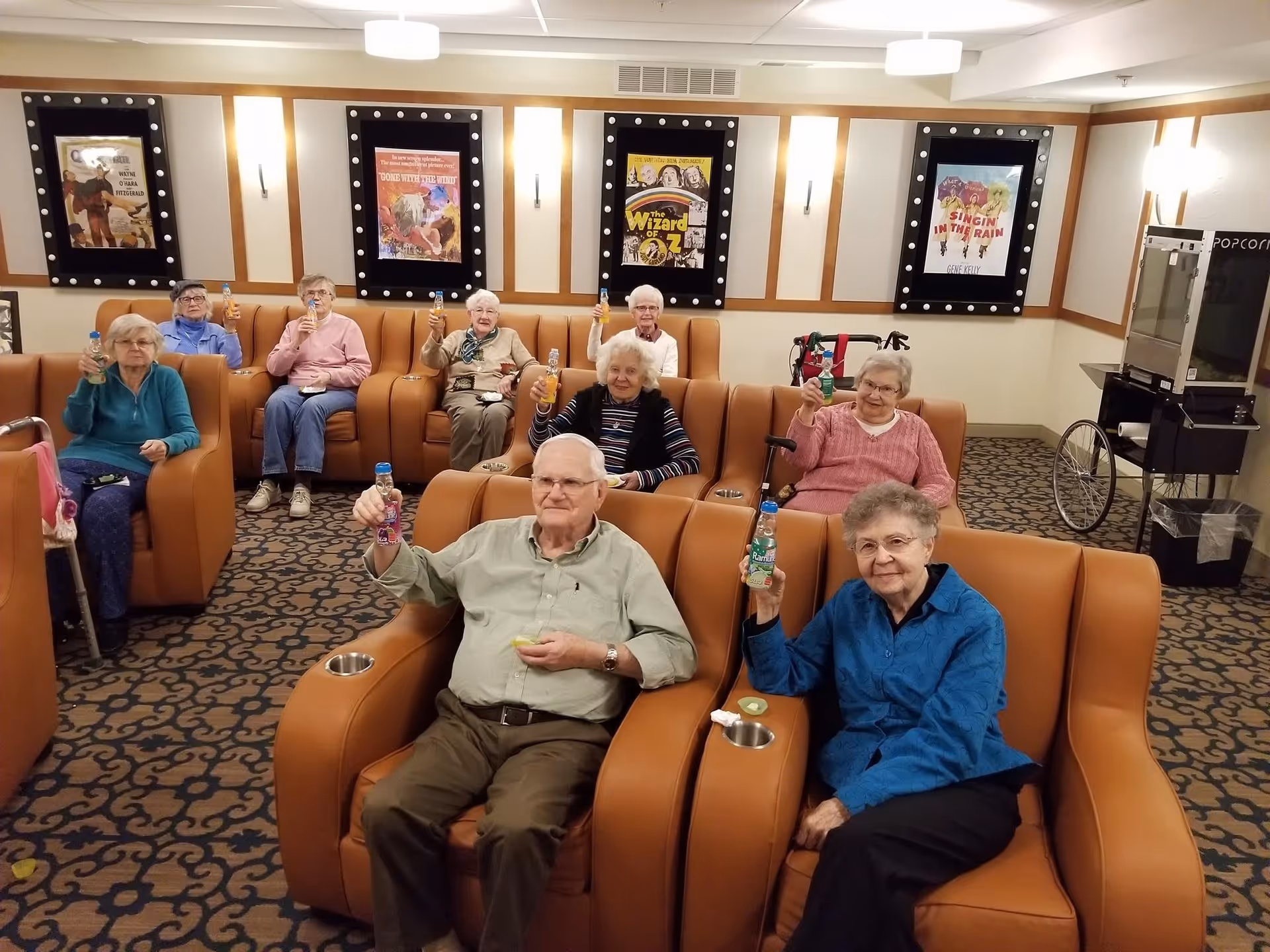 A group of elderly people sitting in comfortable theater-style chairs in a room decorated with classic movie posters on the walls. They are holding up bottles of drinks and smiling, with a popcorn machine visible in the background.