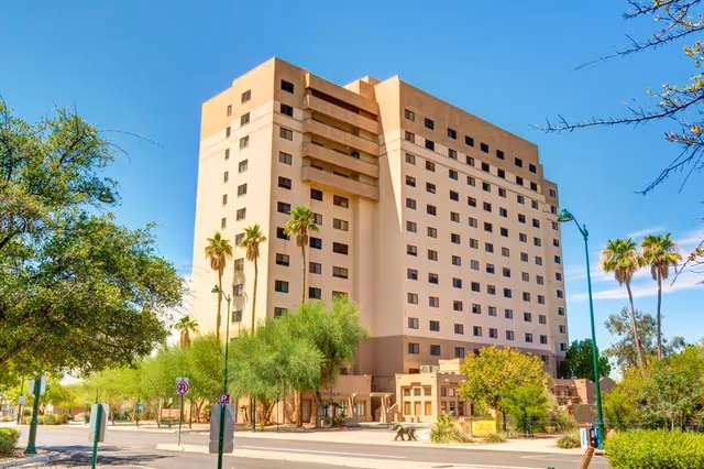 Multi-story beige residential tower with palm trees and a street in front under a clear blue sky.