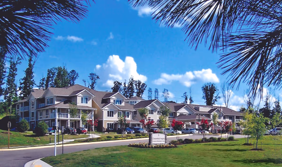 Exterior view of Chesterfield Heights Gracious Retirement Living facility with a large multi-story building, parking lot with cars, green lawn, trees, and a bright blue sky with clouds.