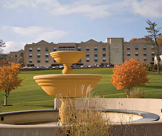 A large beige multi-story building in the background with multiple windows, set behind a green lawn with a few small trees with orange autumn leaves. In the foreground, there is a large yellow tiered fountain with some dry grass around its base under a partly cloudy sky.