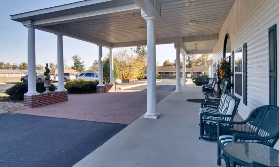 Covered entrance area of a senior living facility with white columns, outdoor seating including chairs with cushions and small tables, and a paved driveway with parked cars in the background.