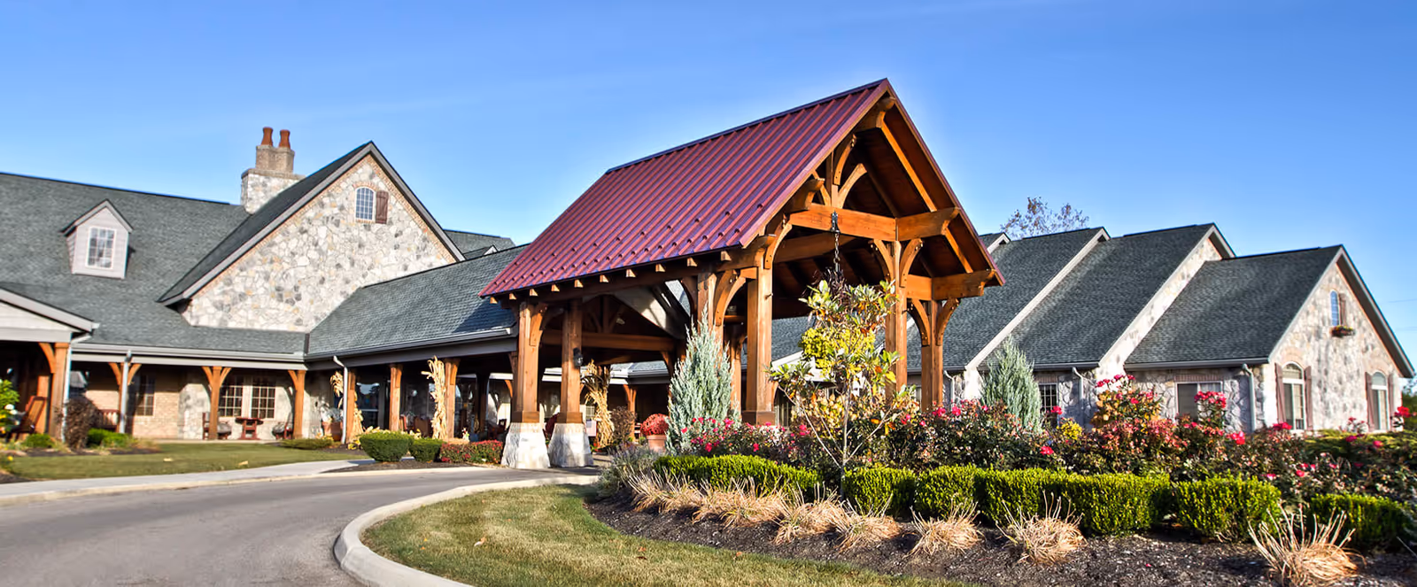 Exterior view of a senior living facility with stone walls and green shingled roofs. The entrance features a wooden covered driveway with a red metal roof. Well-maintained landscaping with bushes, flowers, and grass surrounds the building under a clear blue sky.
