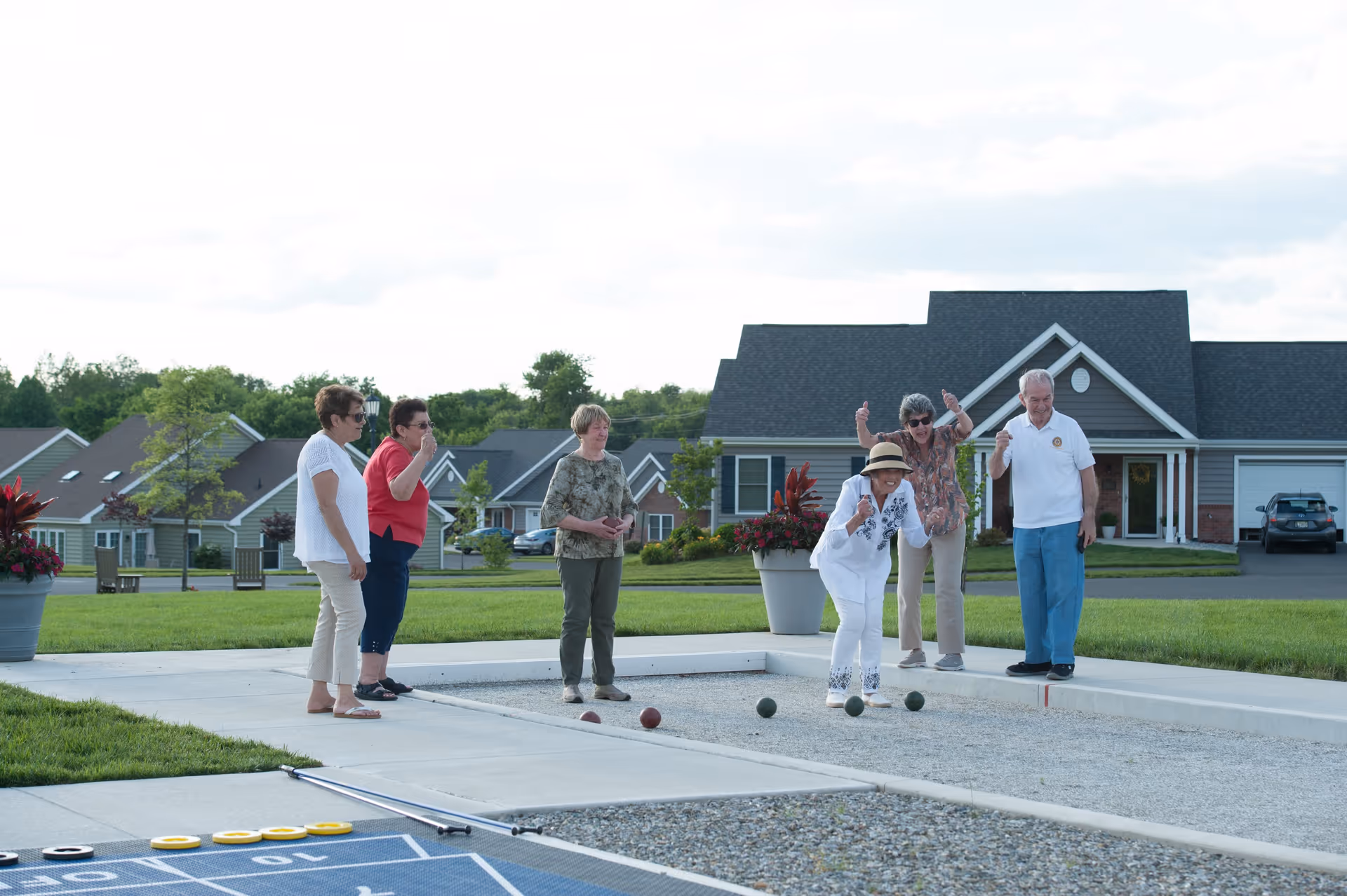 A group of six elderly people playing bocce ball outdoors on a bocce court in a residential community with houses and greenery in the background. One woman is in the act of throwing a bocce ball while others watch and cheer.