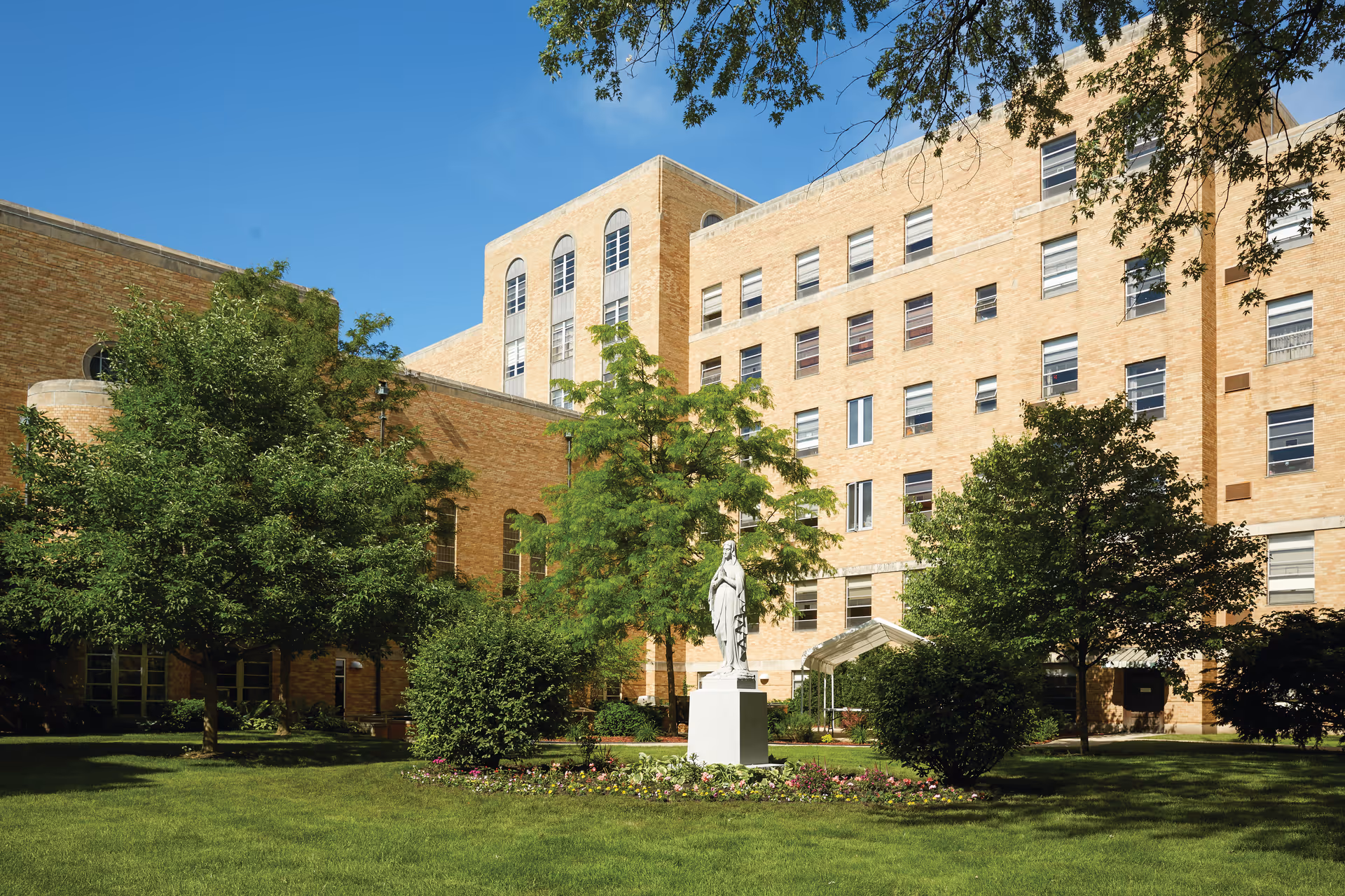 Exterior view of a senior living facility building with multiple stories, surrounded by green trees and a well-maintained lawn. In the center of the lawn is a statue on a pedestal, with flower beds around it. The sky is clear and blue.
