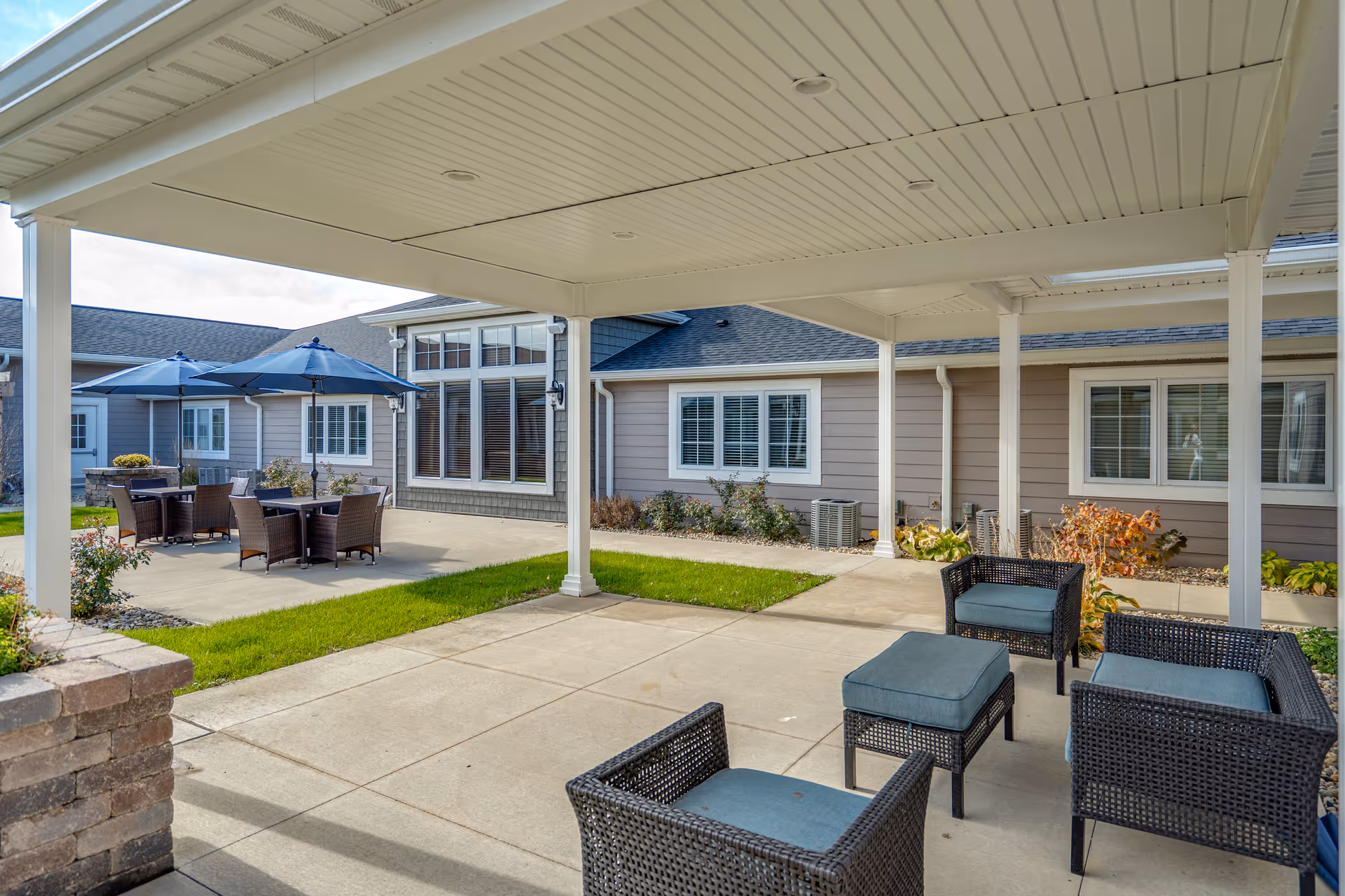 Outdoor patio area at Villas of Holly Brook Assisted Living & Memory Care in Pekin, IL, featuring a covered seating area with wicker chairs and cushions, and an uncovered area with tables, chairs, and blue umbrellas. The building exterior with windows and beige siding is visible in the background.