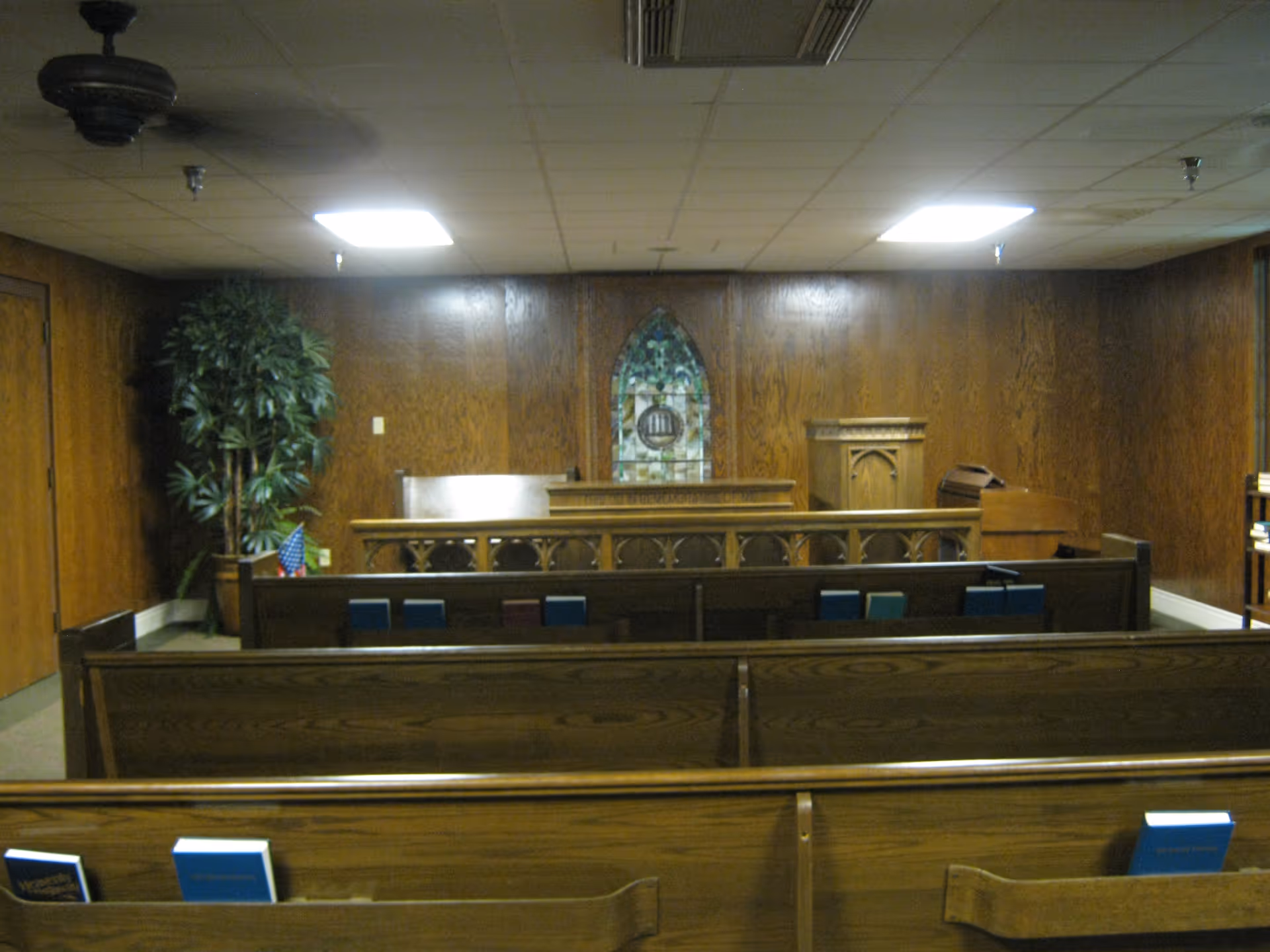 Small wood-paneled chapel interior with wooden pews, a stained-glass window, pulpit, and a potted plant.