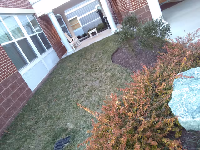Outdoor courtyard area of a facility with a grassy lawn, shrubs, a large rock, and a covered walkway supported by white columns. There are brick walls and windows on the left side, and a bench and chair under the covered area in the background.