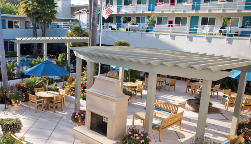 Outdoor patio area at The Village at The Palms featuring wooden tables and chairs with blue umbrellas, a large pergola, a stone fireplace, potted plants, and a multi-story building with balconies in the background.