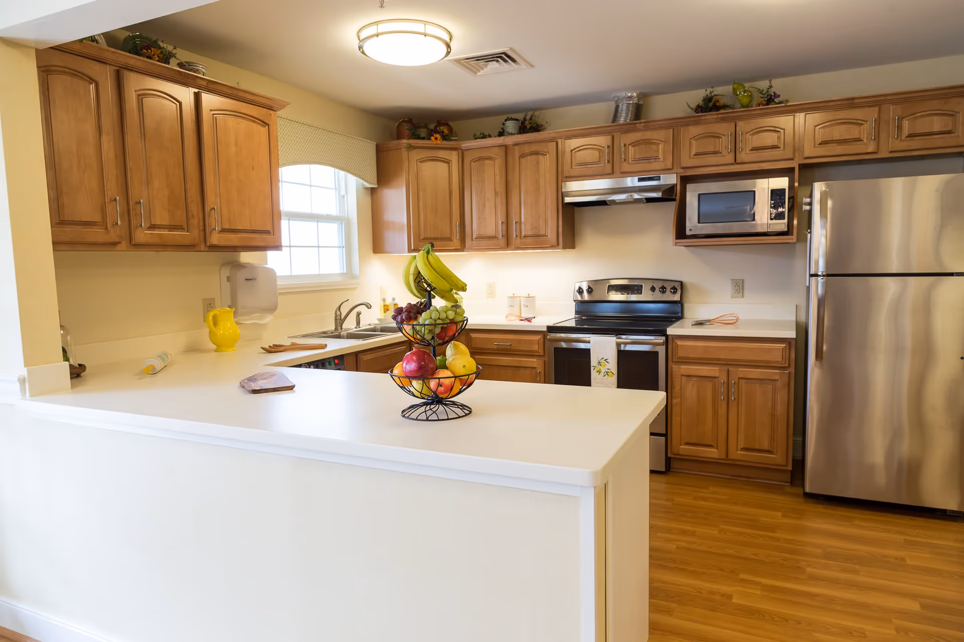 A bright kitchen with wooden cabinets, a stainless steel refrigerator, stove, and microwave. There is a white countertop island with a tiered fruit basket containing bananas, grapes, apples, and pears. A window with a valance lets in natural light, and the floor is wooden.