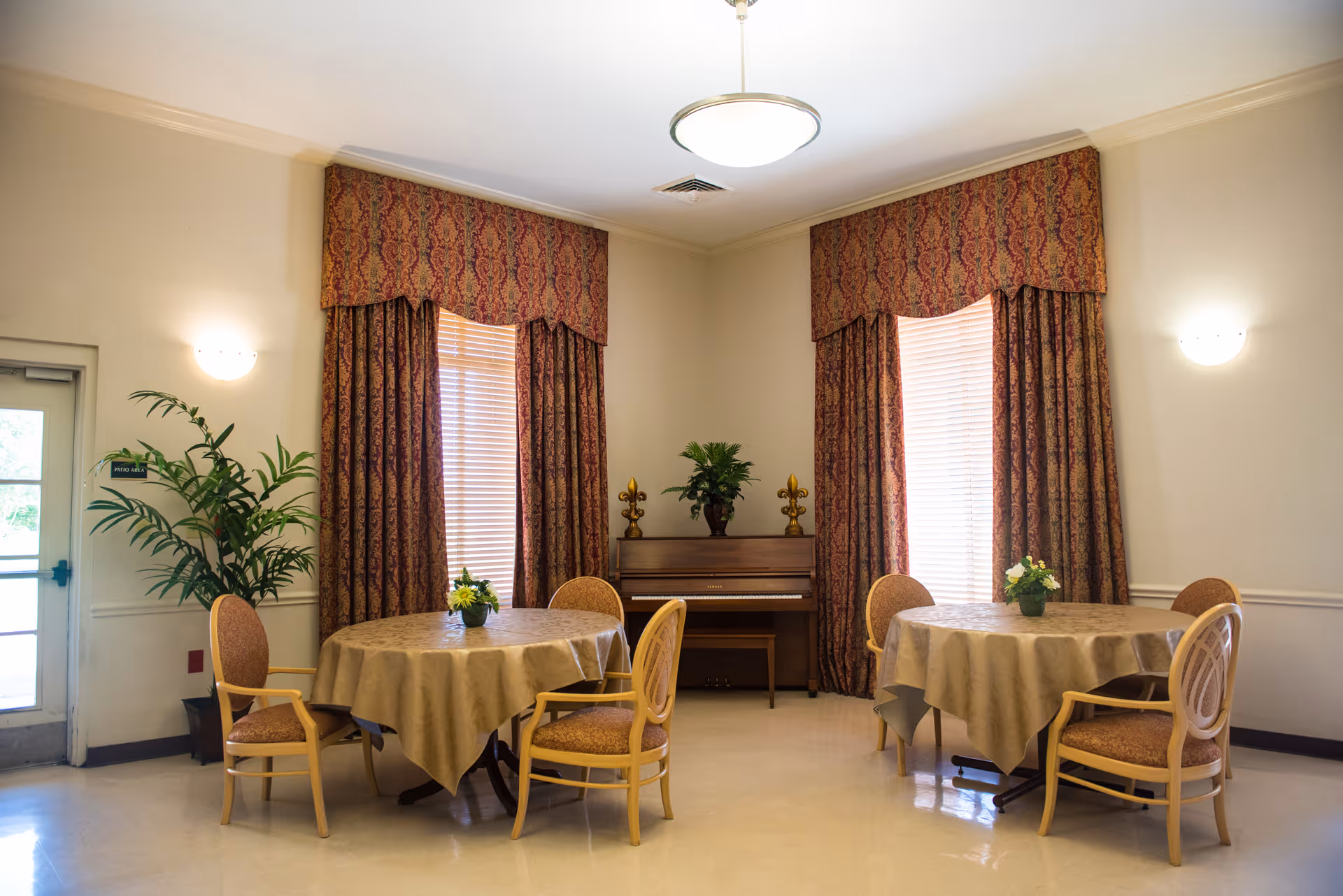 Bright common room with two round tables and chairs, a piano against the back wall, plants, and tall draped windows.
