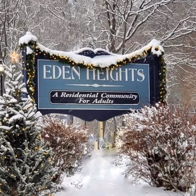 Snow-covered entrance sign reading "Eden Heights: A Residential Community For Adults" surrounded by decorated evergreens and snowy bushes.