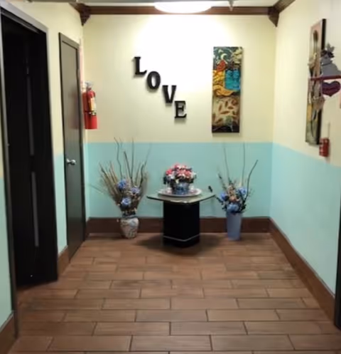 A hallway corner in a facility with light yellow and blue walls, brown tiled floor, and wooden trim. The word 'LOVE' is displayed on the wall with black letters arranged vertically and horizontally. Below the letters is a small black table with a flower arrangement on top, flanked by two vases with decorative branches and flowers. There are doors on the left and right sides of the hallway, a fire extinguisher mounted on the left wall, and decorative wall art on the right.