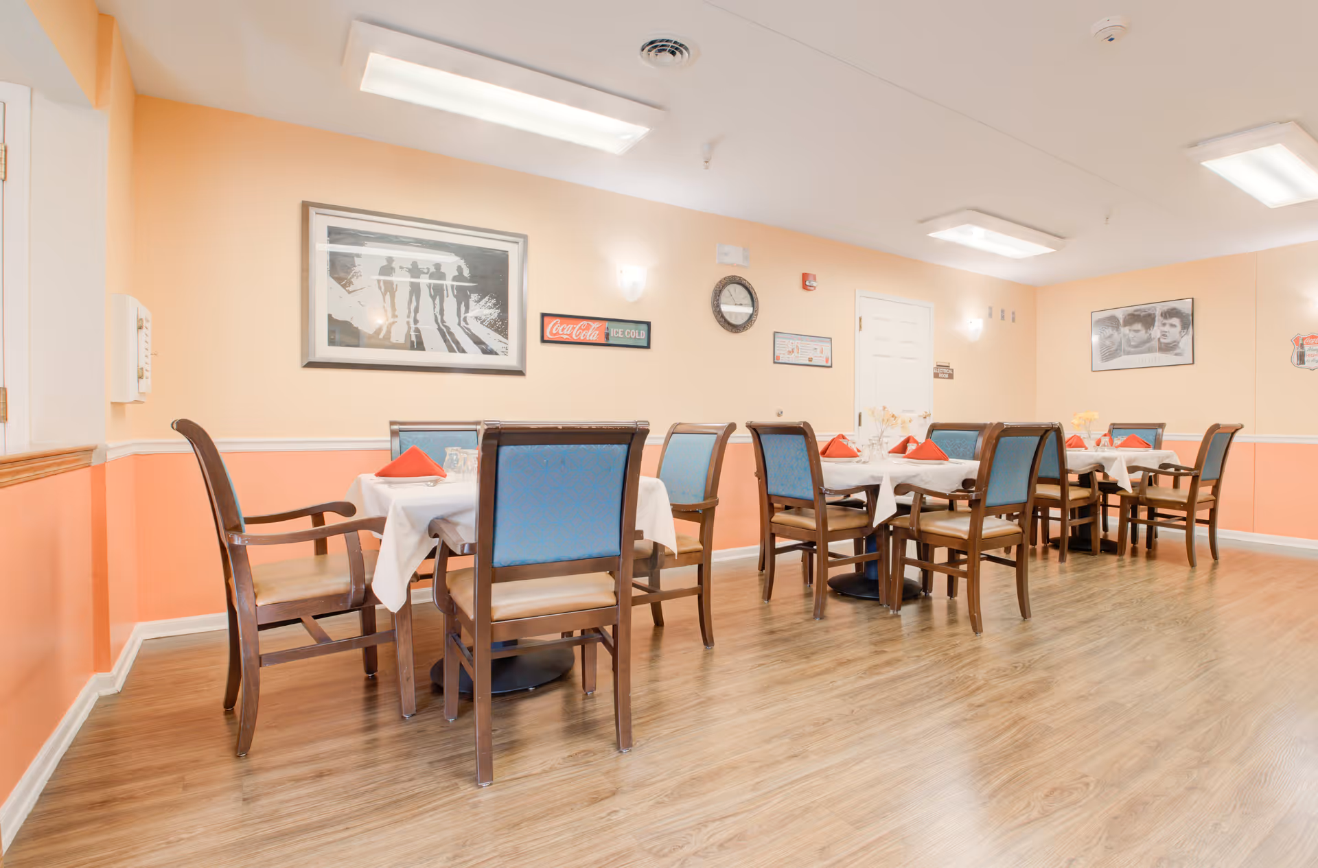 A dining room with wooden floors and peach-colored walls. There are several tables covered with white tablecloths, each set with red napkins and glassware. Wooden chairs with blue cushions surround the tables. The walls are decorated with framed pictures, a clock, and a Coca-Cola sign. The room is well-lit with ceiling lights.