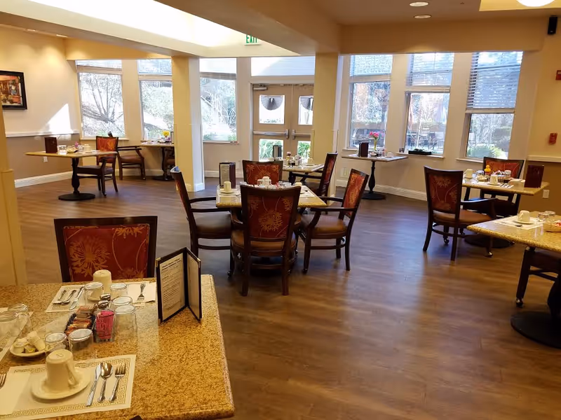 Dining room in an assisted living facility with several set tables and chairs, hardwood floors, and large windows letting in daylight.