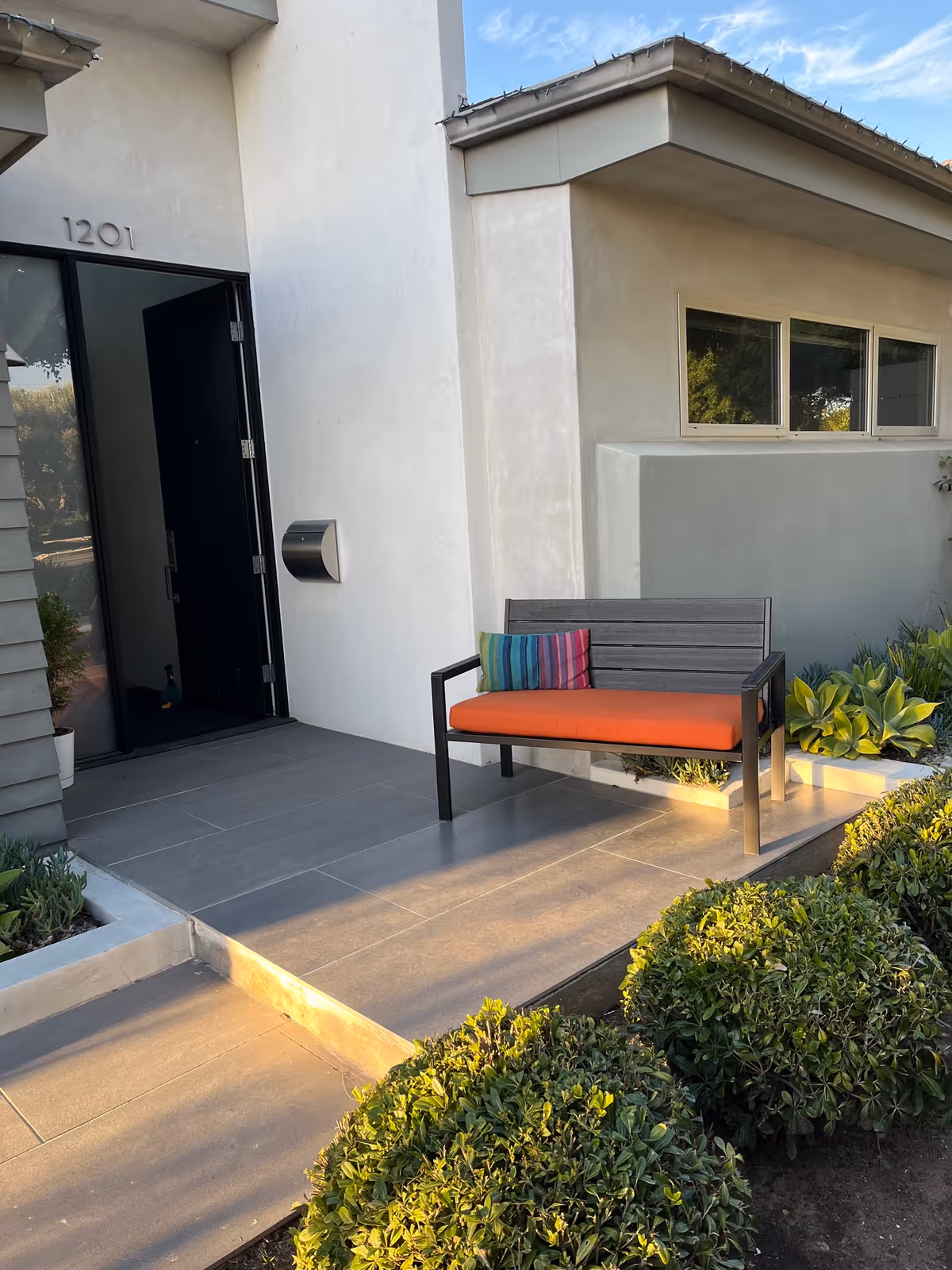 Front entrance of a modern home with a bench that has an orange cushion and striped pillow on a tiled porch, house number 1201 visible.