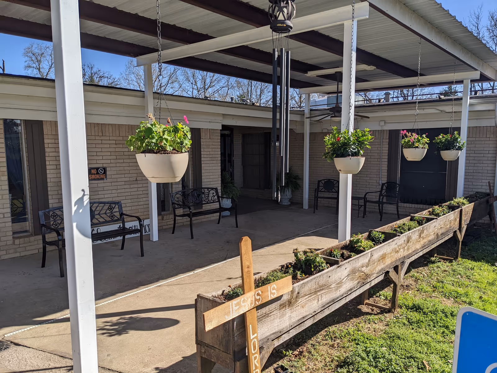 Outdoor covered patio area with hanging flower pots, black metal benches, a wind chime, and a long wooden planter box with small plants. A wooden cross with the words 'JESUS IS LORD' is attached to the planter. The building exterior is visible with beige brick walls and windows.