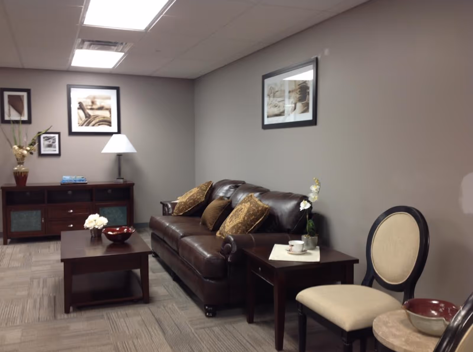 Small common room with a brown leather sofa, coffee table, side chairs and framed artwork on gray walls.
