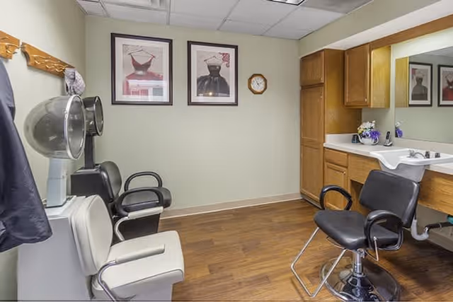 Interior of a hair salon area with three salon chairs, two hair dryers, a large mirror above a countertop with a sink, wooden cabinets, and framed pictures of dresses on the wall.