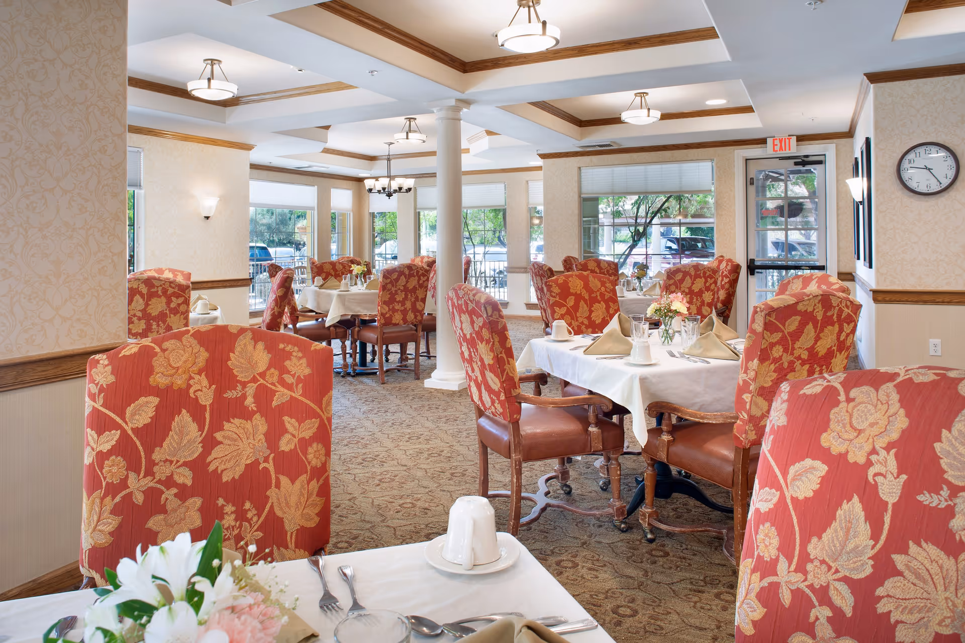 A bright and elegant dining room with tables set for a meal, featuring white tablecloths, folded napkins, glassware, and floral centerpieces. The room has large windows allowing natural light, patterned carpet, and red upholstered chairs with a floral design. Ceiling lights and a clock on the wall are also visible.