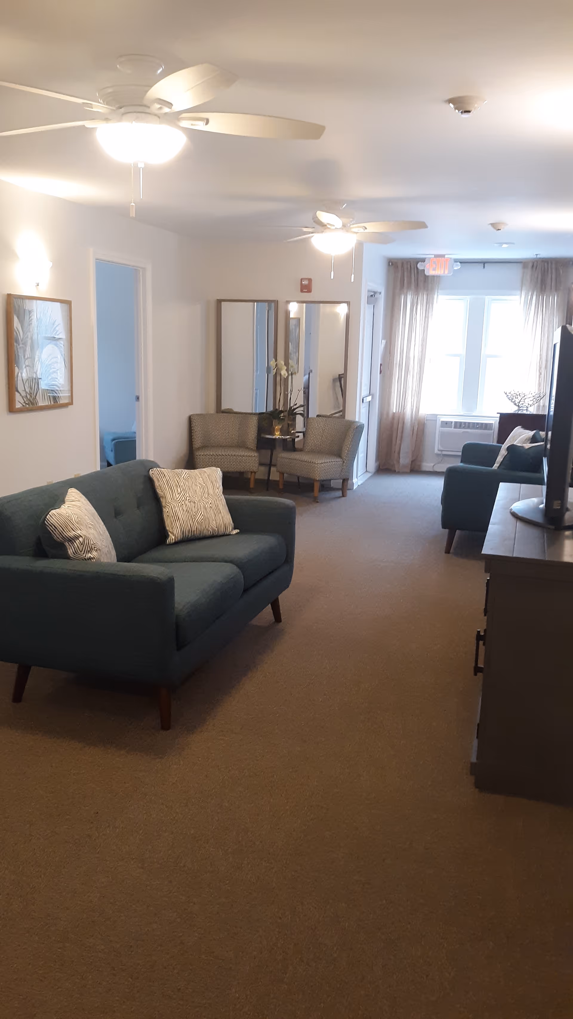 A well-lit living room area with two blue sofas, two patterned armchairs, a TV on a stand, ceiling fans with lights, a framed picture on the wall, and large windows with sheer curtains allowing natural light to enter.