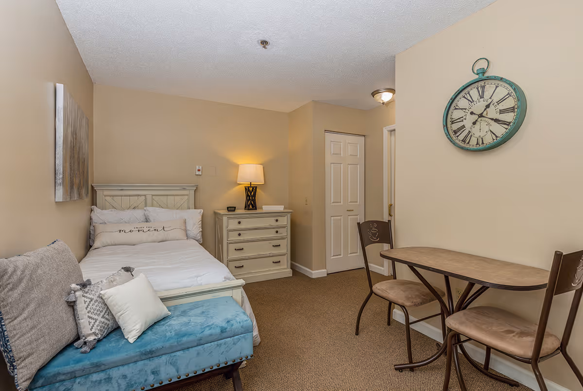 A cozy bedroom in a senior living facility featuring a single bed with white bedding and decorative pillows, a small blue cushioned bench at the foot of the bed, a white dresser with a lamp on top, a small table with two chairs, and a large vintage-style wall clock. The walls are painted beige and the floor is carpeted.