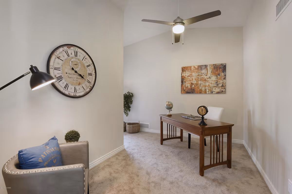 A small office or study room with beige walls and carpet. There is a wooden desk with a globe, a small clock, and an open book on it. A white chair is placed behind the desk. On the left side, there is a gray armchair with a blue pillow and a black floor lamp. A large vintage-style wall clock hangs above the armchair. A ceiling fan with a light is mounted on the ceiling, and an abstract painting is hung on the wall behind the desk. A plant and a basket are visible in the corner.