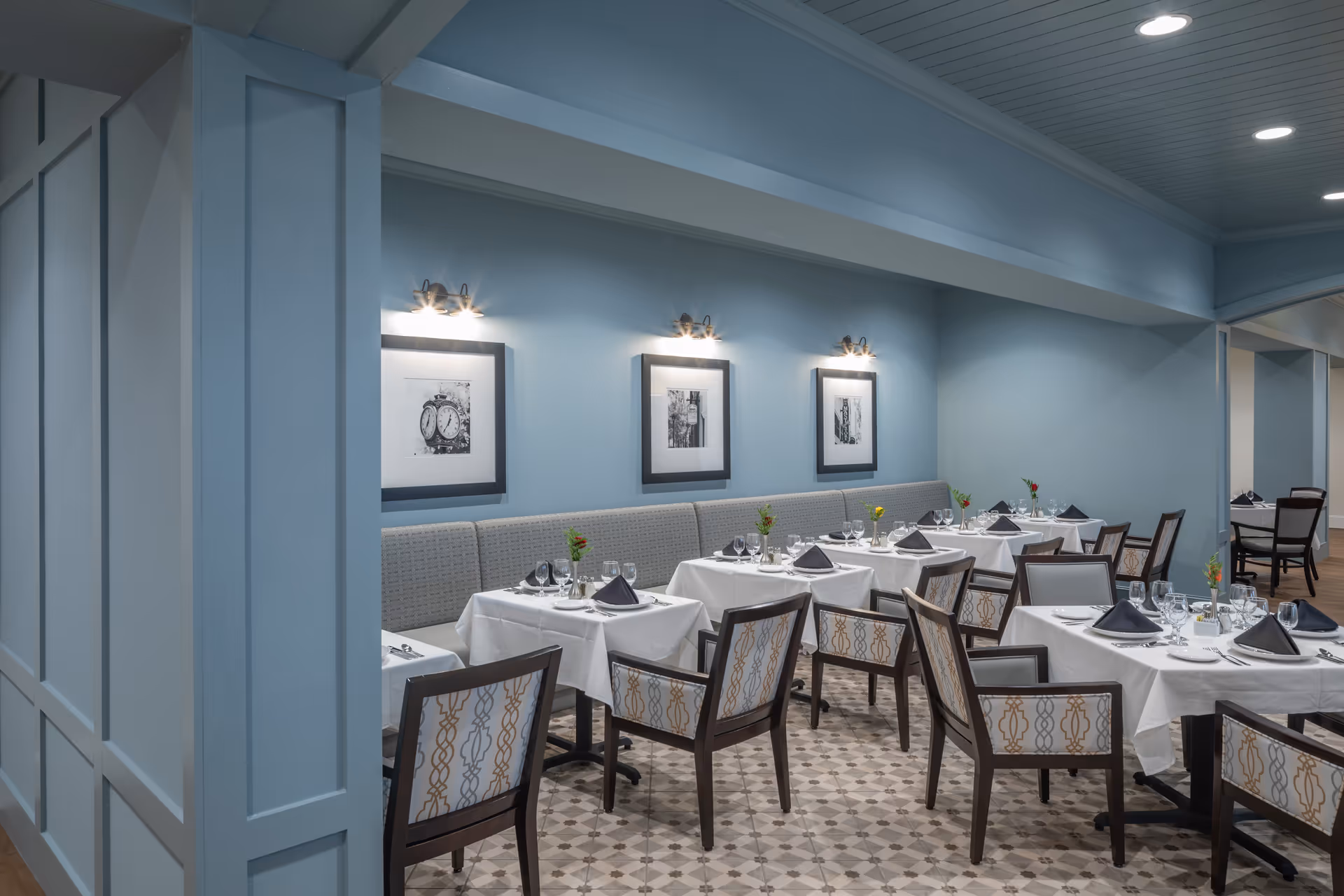 A dining room with several tables covered in white tablecloths, each set with plates, glasses, silverware, and black napkins. The chairs have patterned upholstery and there is a long cushioned bench along the blue wall. Three black and white framed pictures are hung on the wall above the bench, each illuminated by a wall-mounted light fixture. The floor has a patterned tile design and the ceiling has recessed lighting.