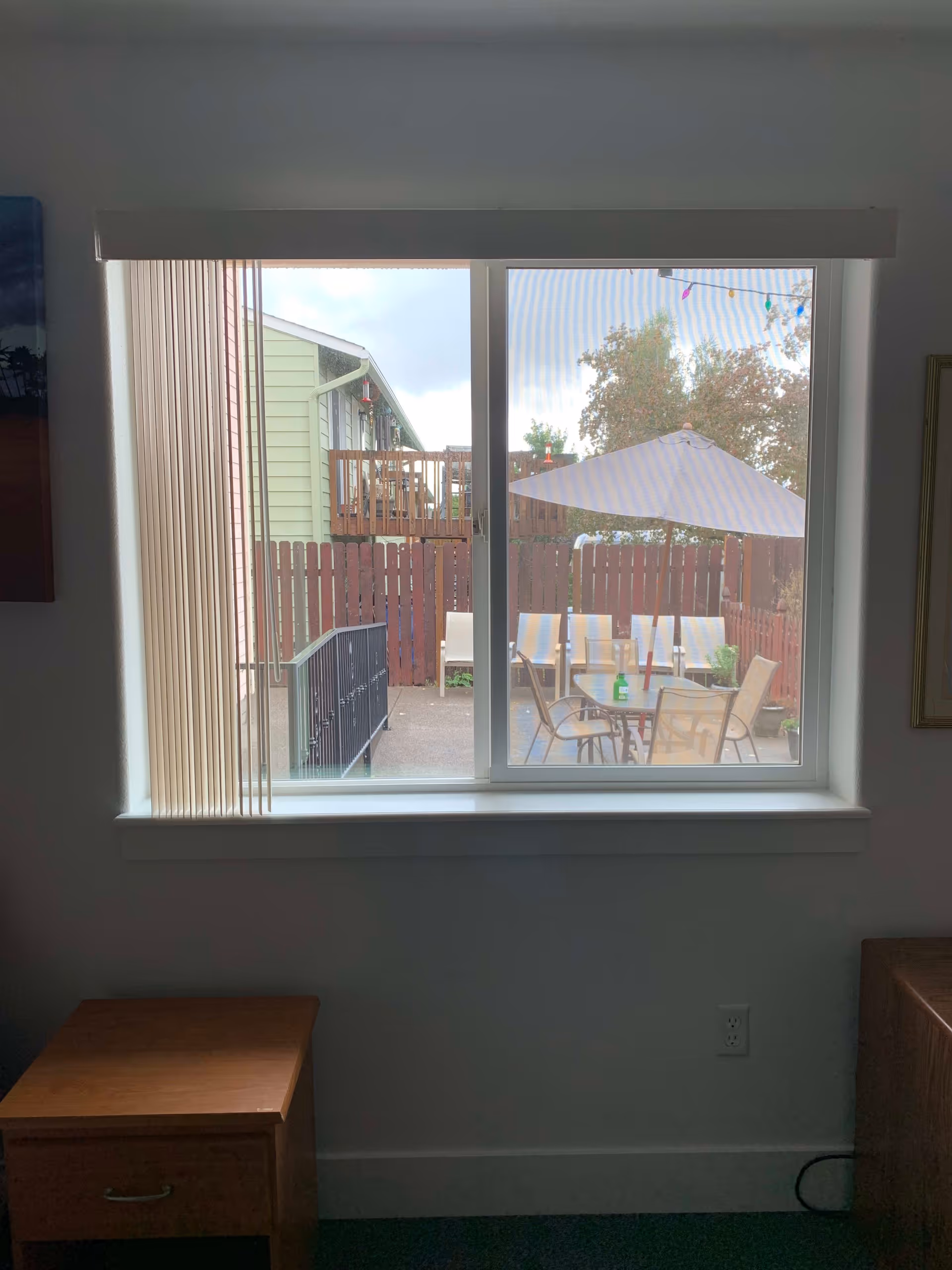 View through a room window onto a fenced outdoor patio with a table, chairs, and an umbrella.