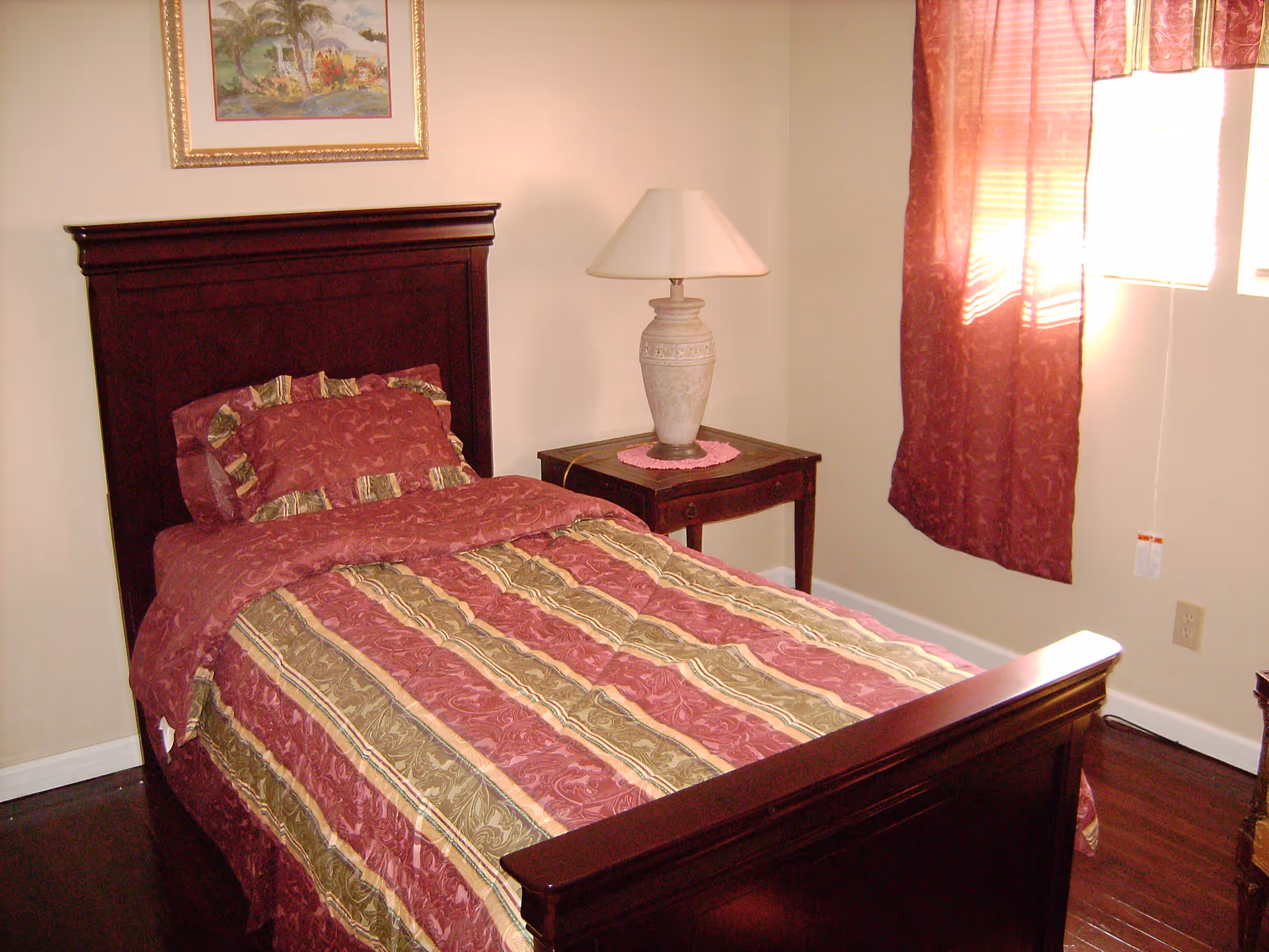 A bedroom with a single bed featuring a dark wooden headboard and footboard. The bed is made with a red, gold, and green patterned comforter and matching pillow. Next to the bed is a wooden nightstand with a decorative lamp on top. A framed painting hangs on the wall above the headboard, and a window with red curtains allows sunlight to enter the room.