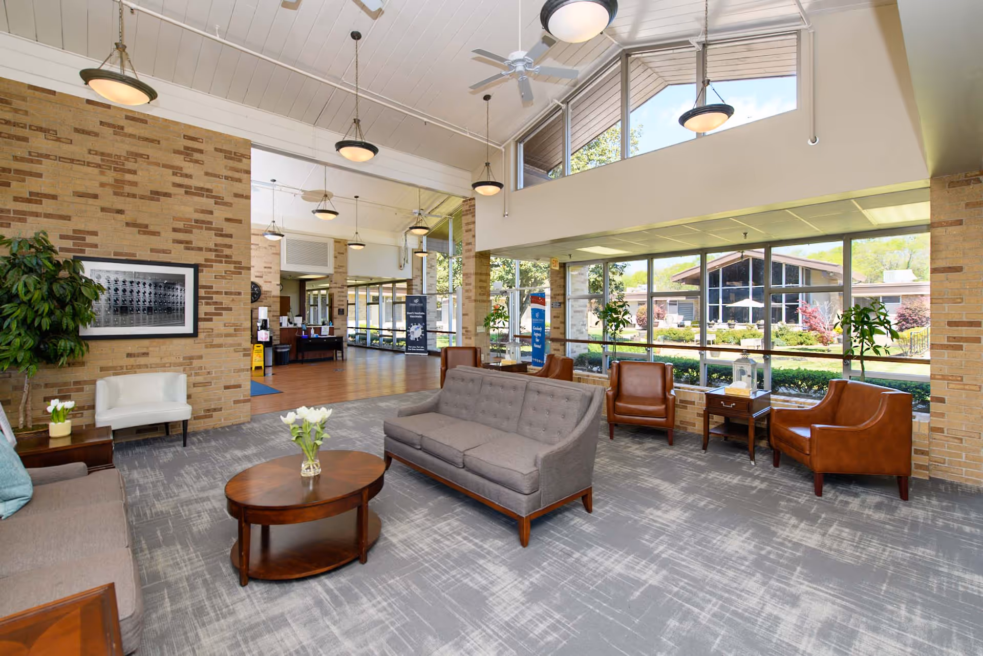 A spacious and well-lit living room area in a senior living facility with large windows allowing natural light to fill the space. The room features a gray sofa, brown leather armchairs, a wooden coffee table with a vase of white flowers, and a side table with a plant. The walls are made of light brown brick, and there are ceiling fans and hanging lights. Outside the windows, a garden and another building are visible.