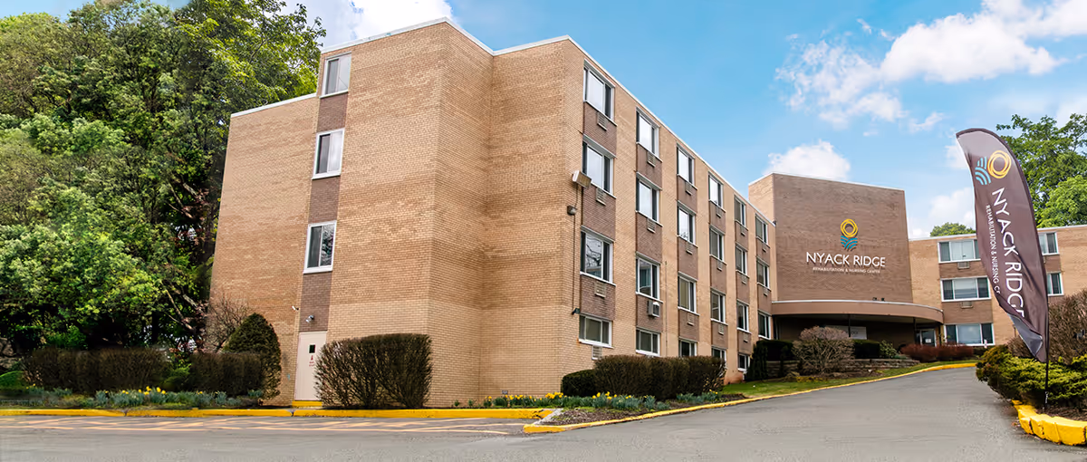 The front exterior of Nyack Ridge Rehabilitation & Nursing Center, a multi-story brick building with a driveway, landscaped shrubs, and a flag bearing the facility name.
