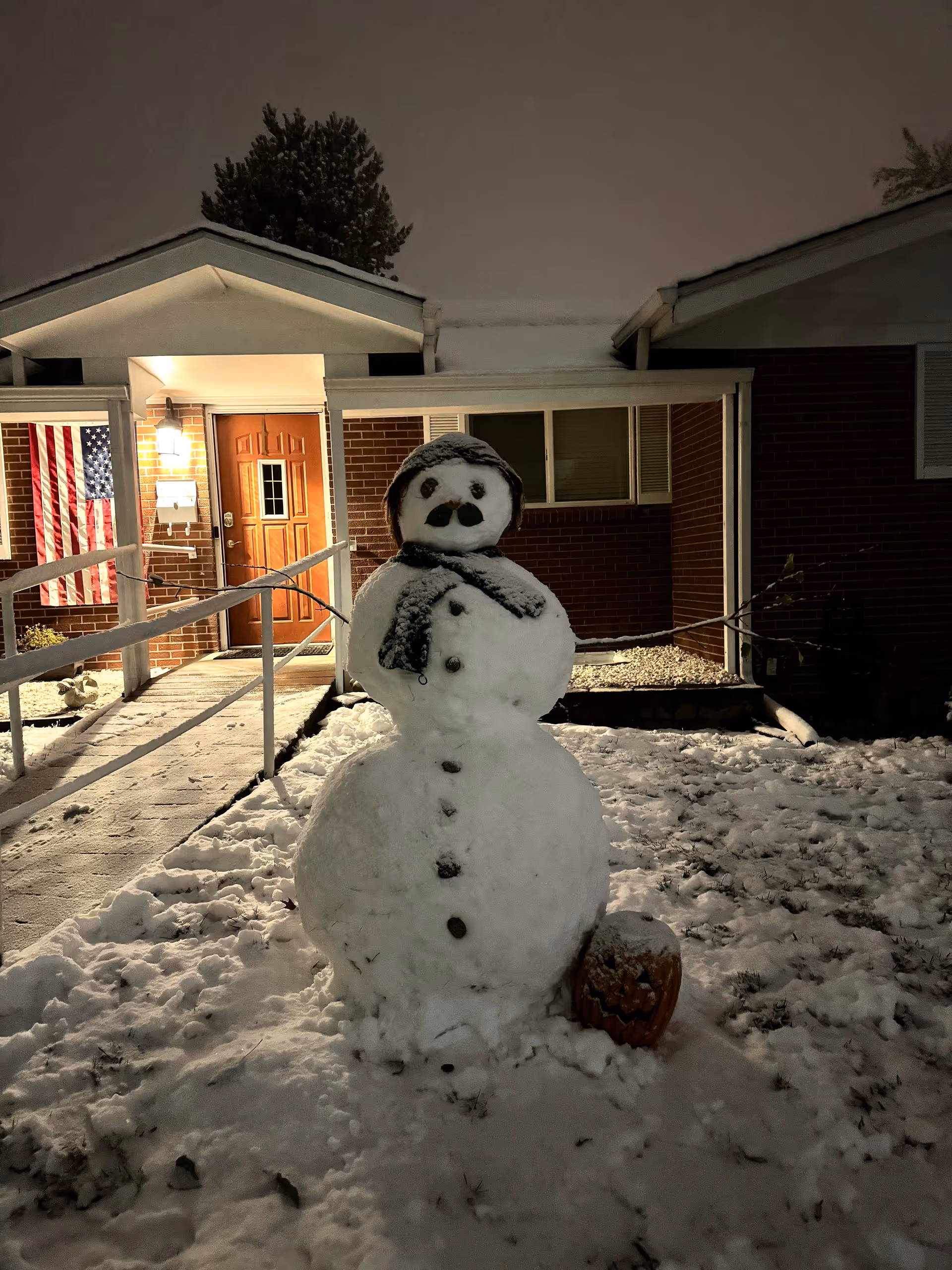 A snowman with a scarf and hat stands in front of a brick building entrance at night. The building has a lit porch light, a wooden door, and an American flag hanging beside the door. Snow covers the ground and a carved pumpkin is placed near the snowman.