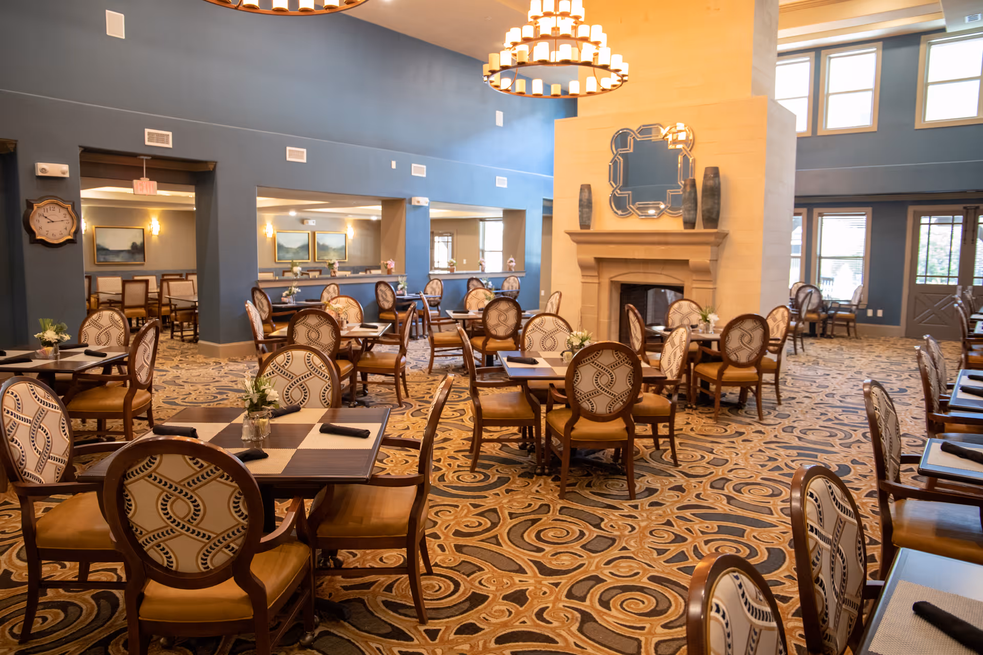 A spacious dining room with multiple wooden tables and patterned chairs arranged neatly. The room features a large fireplace with decorative vases and a mirror above it. The walls are painted blue, and the floor is covered with a patterned carpet. Large windows allow natural light to brighten the space, and elegant chandeliers hang from the ceiling.