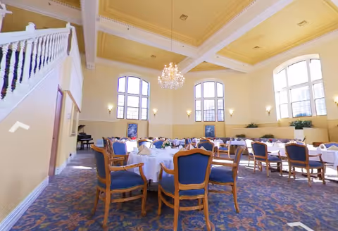 A spacious dining room with multiple round tables covered with white tablecloths and set with napkins and centerpieces. The room features large arched windows, a chandelier hanging from a high ceiling with decorative beams, wall sconces, and a carpeted floor. There is a staircase on the left side and a piano in the back corner.