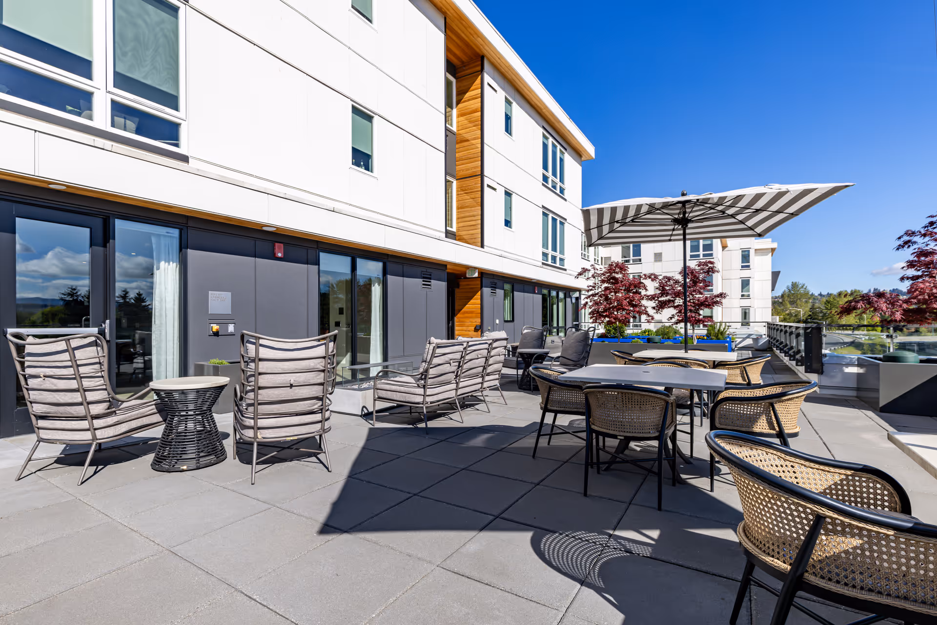 Outdoor patio area at Merrill Gardens at Tukwila featuring multiple seating arrangements including cushioned chairs, wicker chairs around tables, and a large striped umbrella. The patio is adjacent to a modern building with large windows and a clear blue sky overhead.