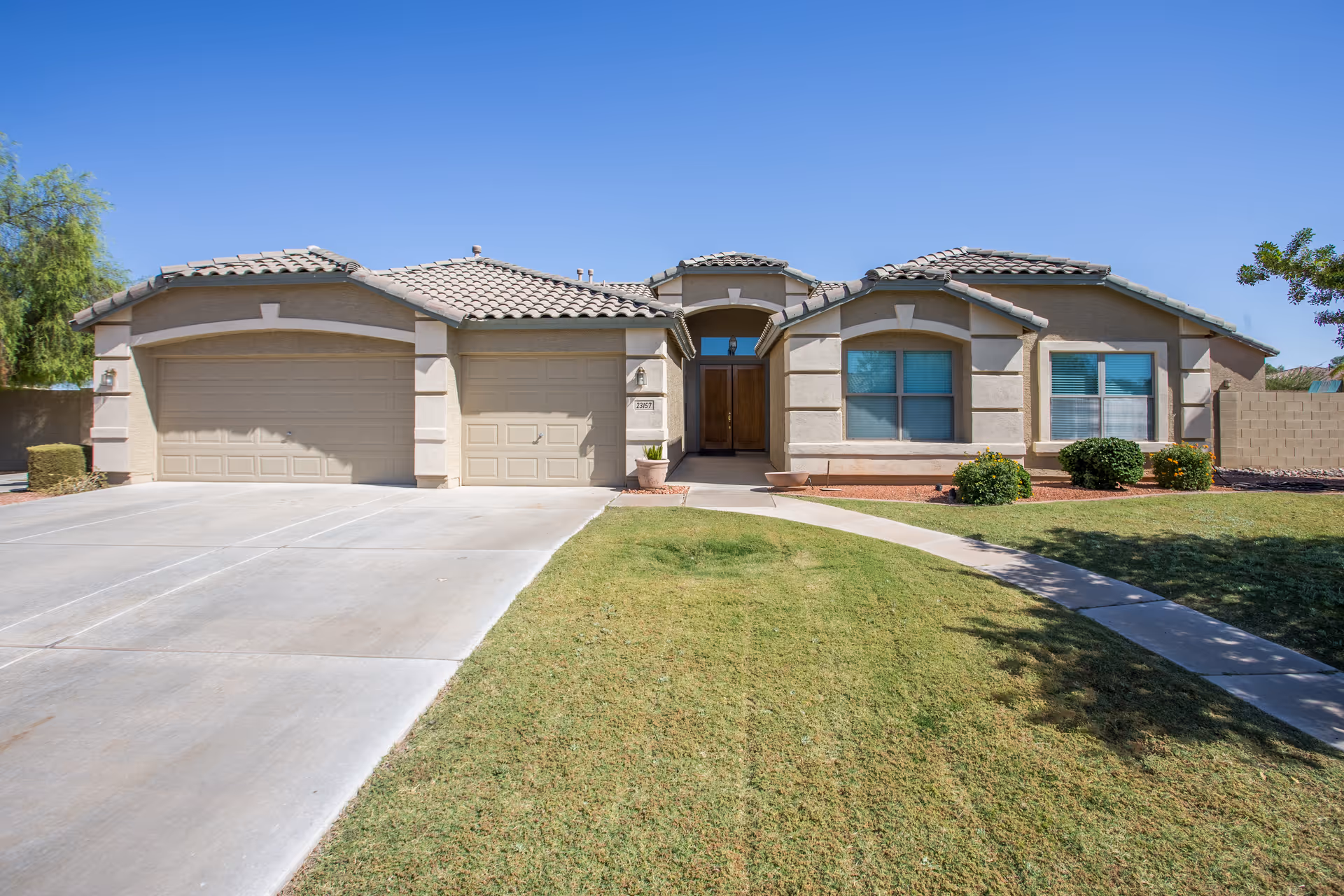 Front exterior view of a single-story residential building with a tiled roof, three garage doors, a concrete driveway, a walkway, and a well-maintained lawn with bushes under a clear blue sky.