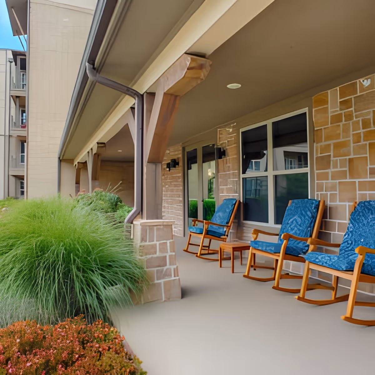 Covered outdoor patio area at Discovery Village at Alliance Town Center with wooden rocking chairs featuring blue cushions, stone pillars, and lush green plants and shrubs along the edge.