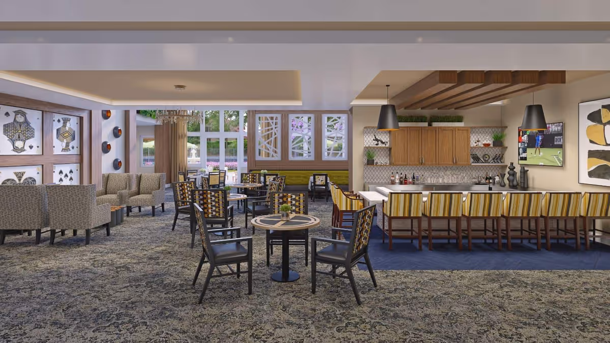 Open communal dining and lounge area with multiple tables and chairs, a long striped bar with stools, and a kitchenette and TV in the background.
