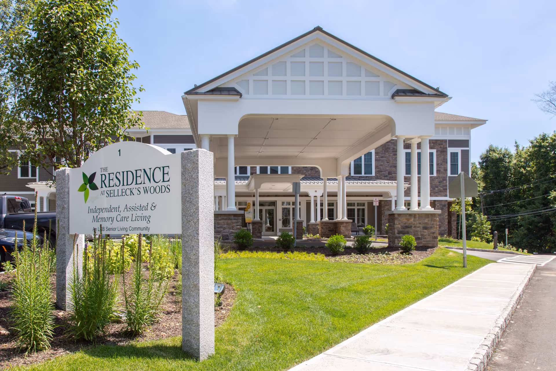 Front exterior view of The Residence at Selleck's Woods senior living facility with a covered entrance, stone pillars, and a sign indicating independent, assisted, and memory care living. The building is surrounded by green landscaping and a clear blue sky.