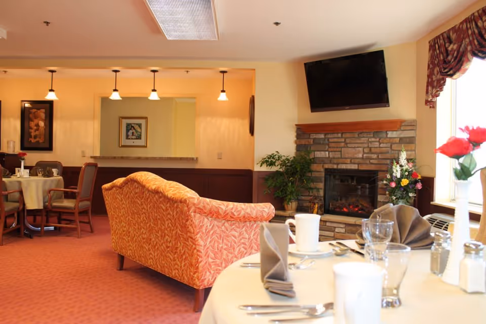 A cozy common area in a senior living facility featuring a patterned orange couch facing a stone fireplace with a mounted flat-screen TV above it. There are tables set with napkins, cups, glasses, and silverware, along with floral centerpieces. The room has warm lighting with hanging pendant lights and framed artwork on the walls.