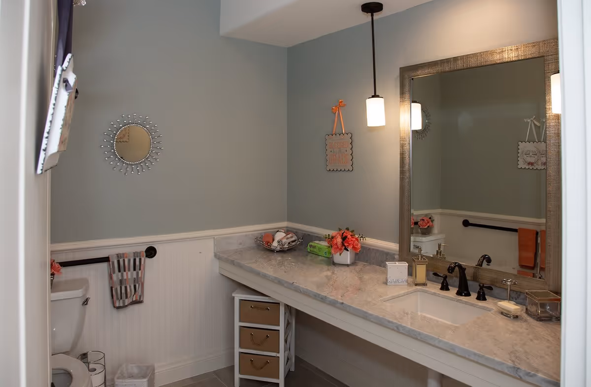 A clean and well-lit bathroom with a toilet, a large marble countertop with a sink, a large framed mirror, a small decorative round mirror on the wall, a towel hanging on a black towel rack, a small set of drawers under the counter, and a vase with pink flowers on the countertop.