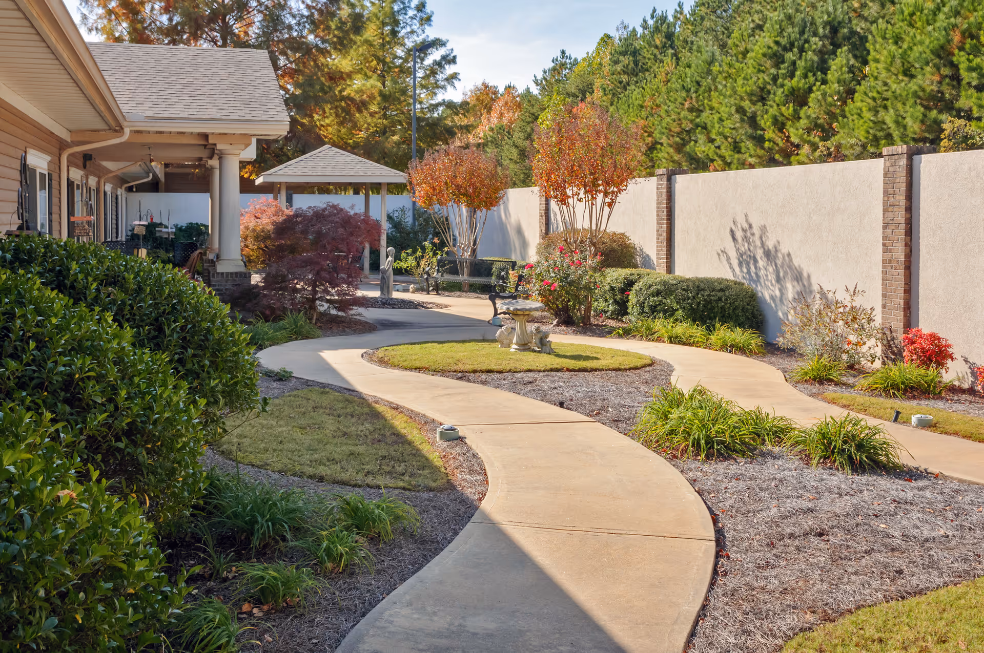 Curved concrete walkway through a landscaped courtyard with a gazebo, benches, and shrubs beside a building.