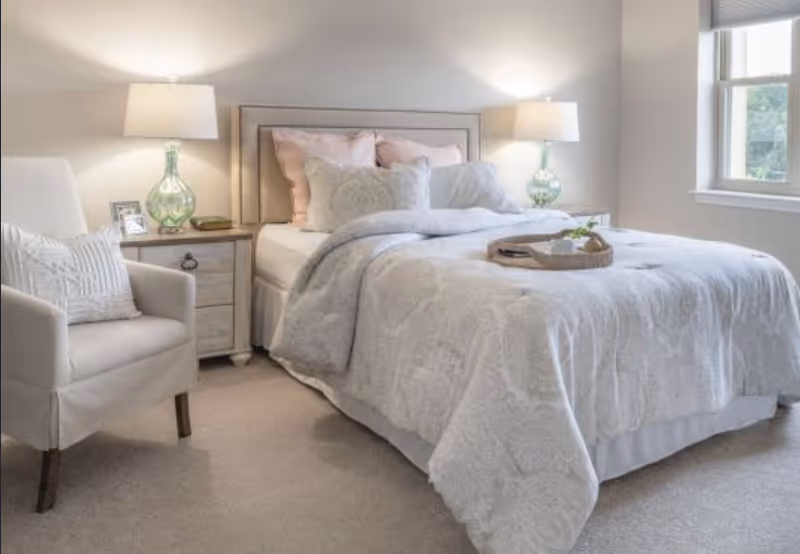 A neatly made bed with light-colored bedding and multiple pillows in a bright bedroom. There is a beige armchair with a decorative pillow to the left of the bed, and two bedside tables each with a lamp. A window on the right side lets in natural light.