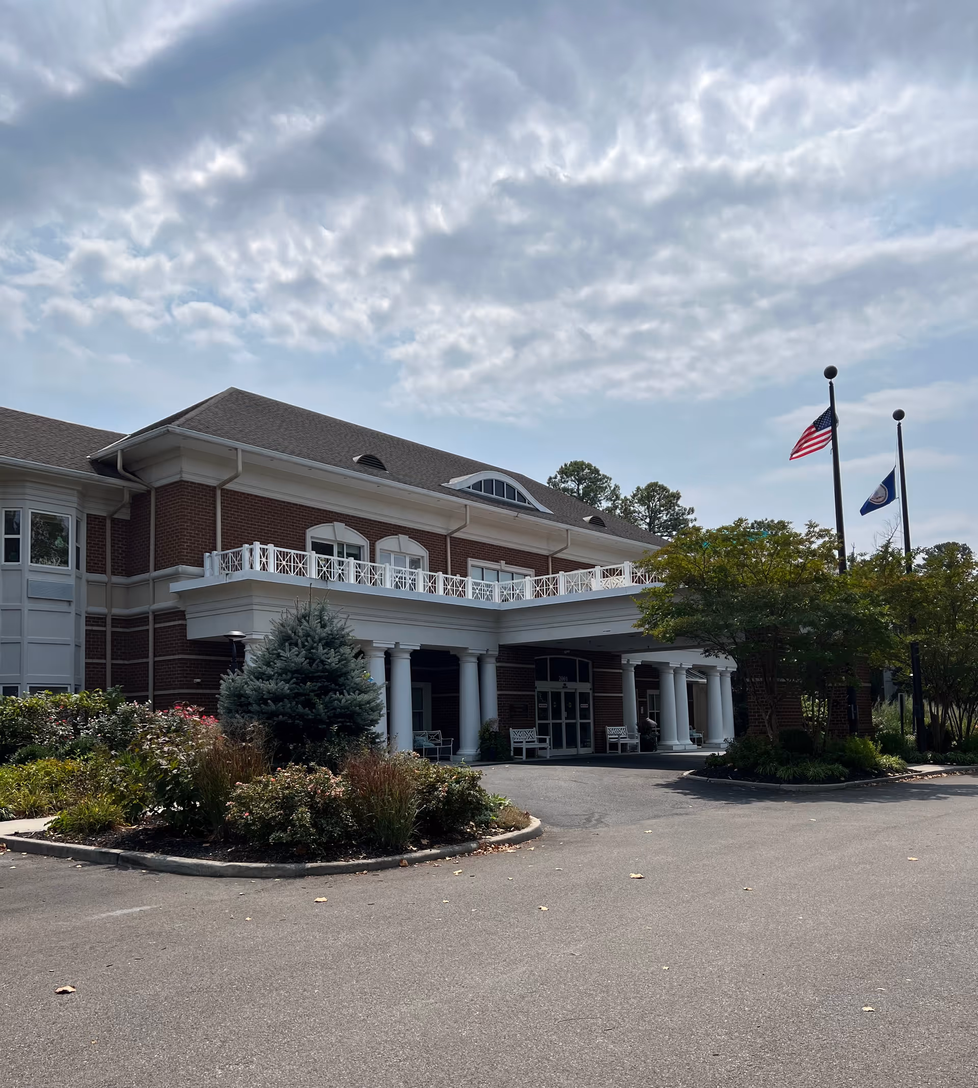 Front exterior view of a senior assisted living facility building with brick walls, white columns, a covered entrance, and a balcony above. There are two flagpoles with the American flag and another flag, surrounded by landscaped bushes and trees under a partly cloudy sky.
