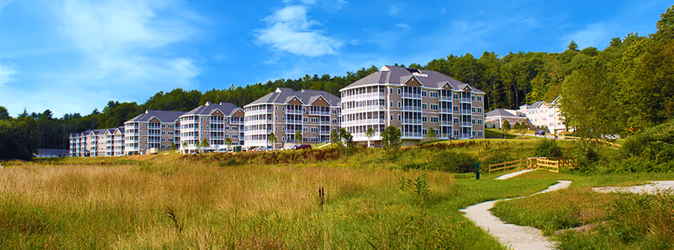 A large senior living facility with multiple connected buildings surrounded by green grass, trees, and a walking path under a blue sky with some clouds.
